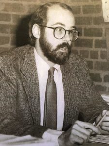 A young man with pale skin, round glasses, and a dark beard sits at a desk in front of brick wall, speaking to someone off-camera. He is wearing a tweed jacket over a button down and tie.