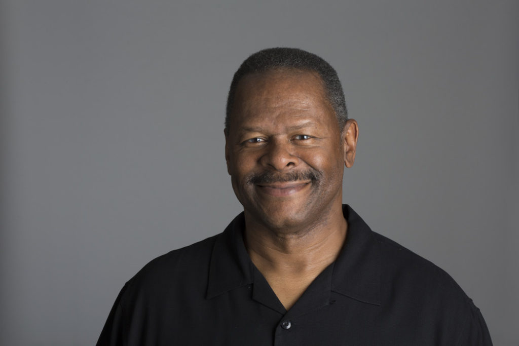 A headshot of Curtis J. Moody, a middle-aged man with brown skin, close-cropped salt-and-pepper hair, and a mustache. He is wearing a black polo, standing against a gray background, and smiling.