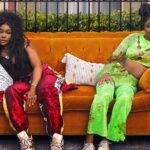 Two young women frustratedly sit on their couch, which has been moved to the curb by their landlord.